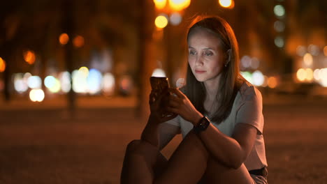 A-young-woman-looks-into-the-smartphone-and-writes-text-messages-on-the-Internet-against-the-backdrop-of-the-night-city.-Girl-businessman-working-on-vacation-remote-work-via-mobile-phone.-Gadget-addiction